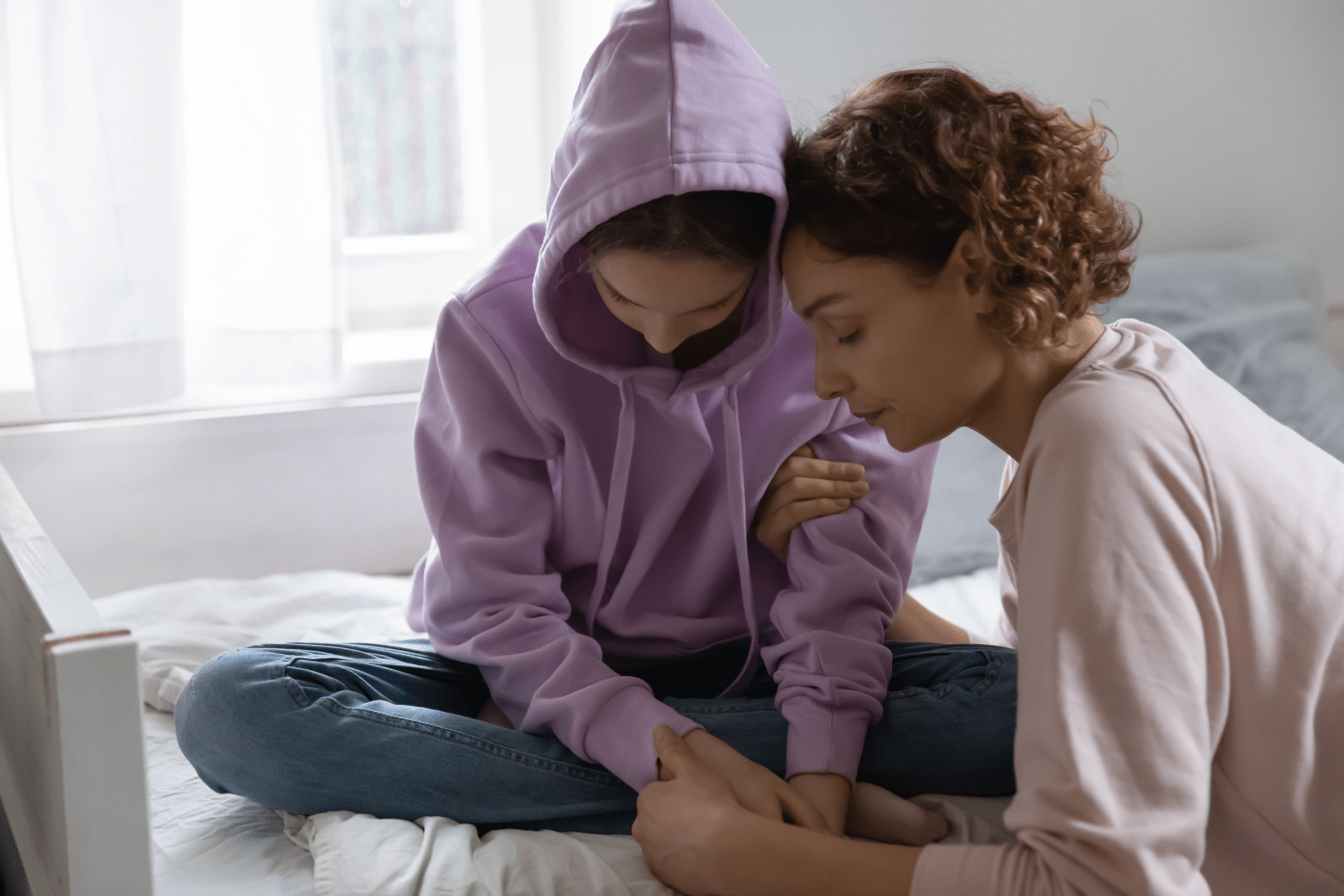 A mother consoles her daughter, who sits cross-legged in blue jeans and a purple sweater with her head down in a depressed state